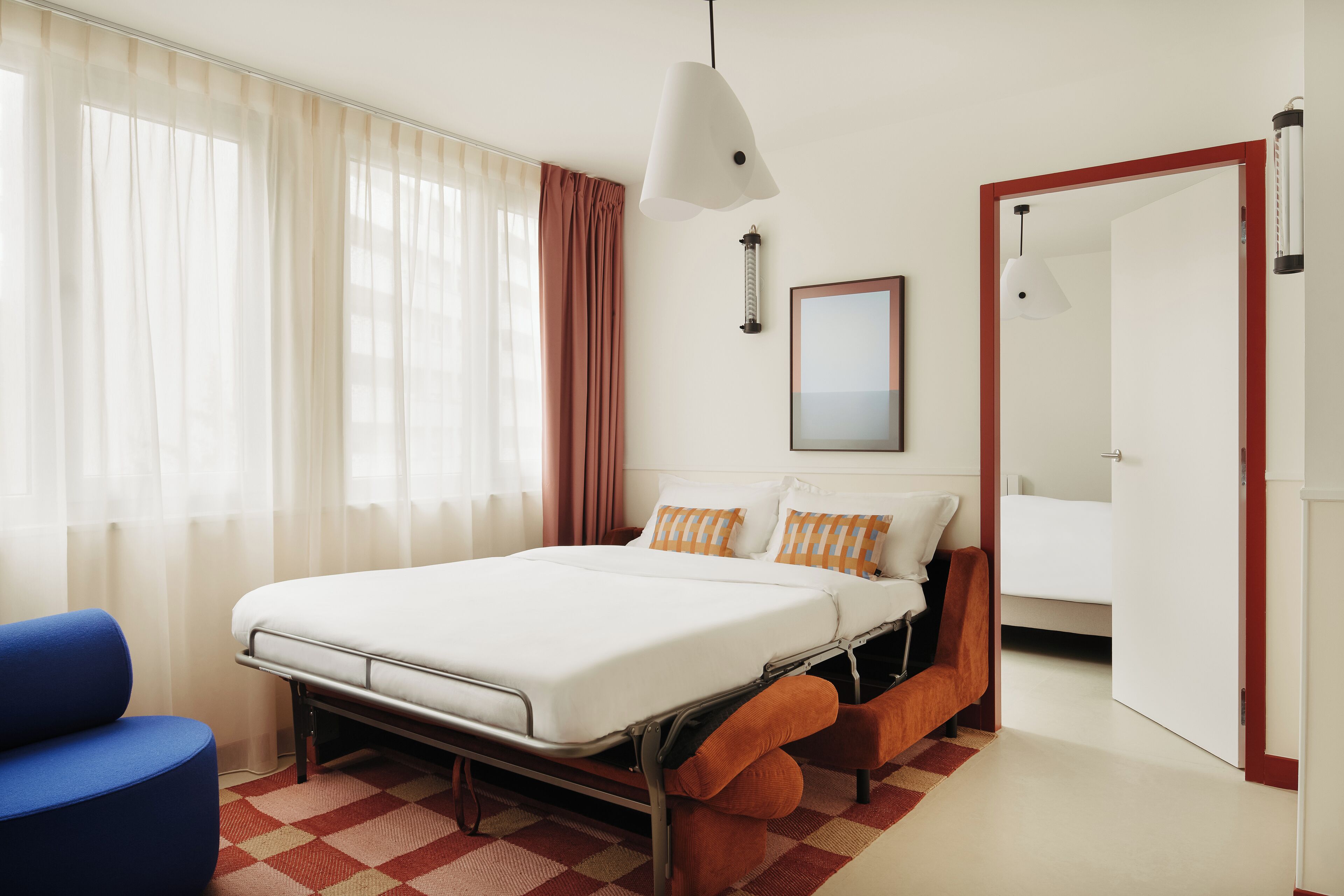 A modern hotel room with a large bed, a blue armchair, and a red and white checkered rug.