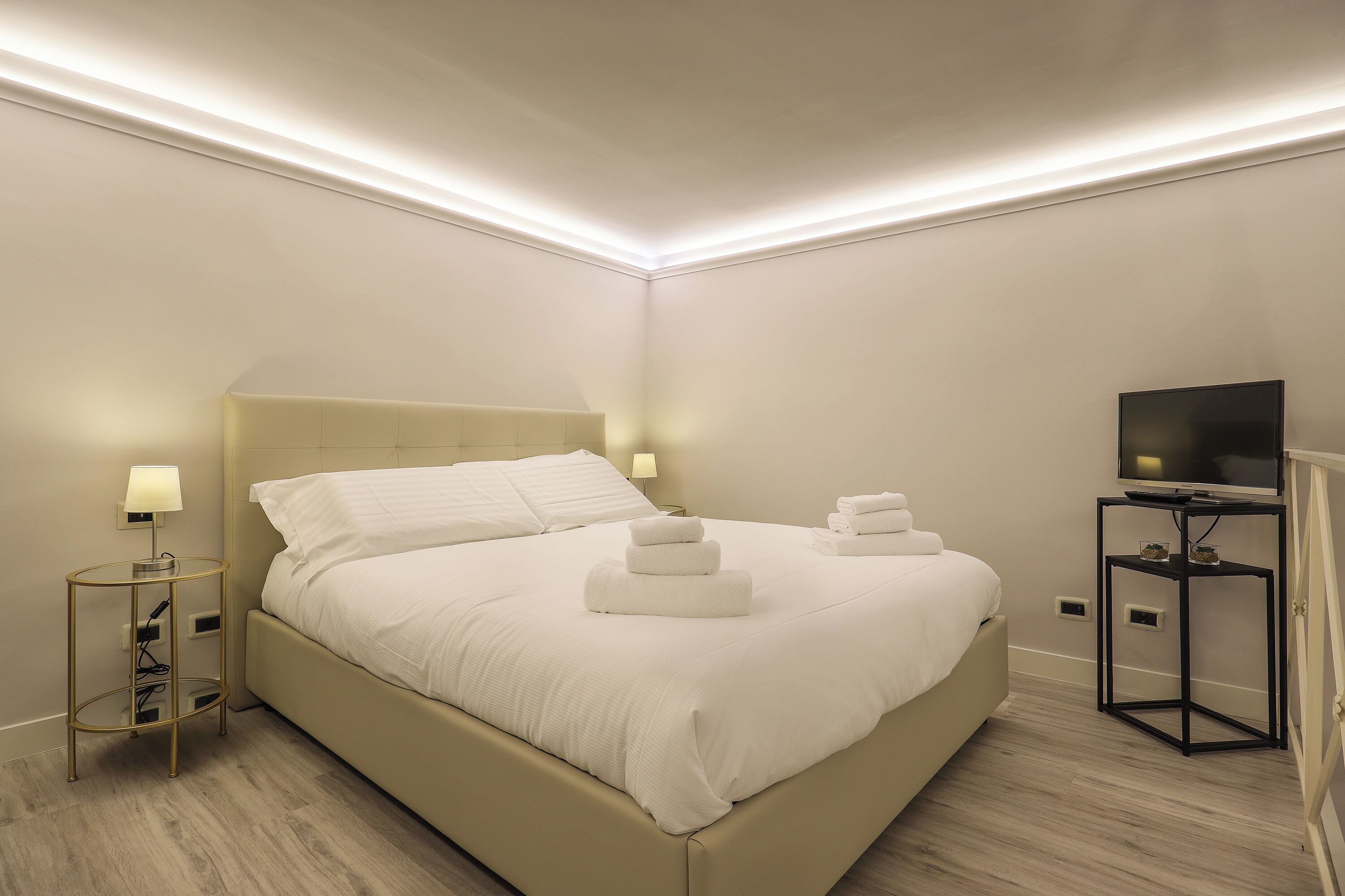 A modern hotel room with a large bed, bedside lamps, a TV on a stand, and a flat-screen TV on the wall.
