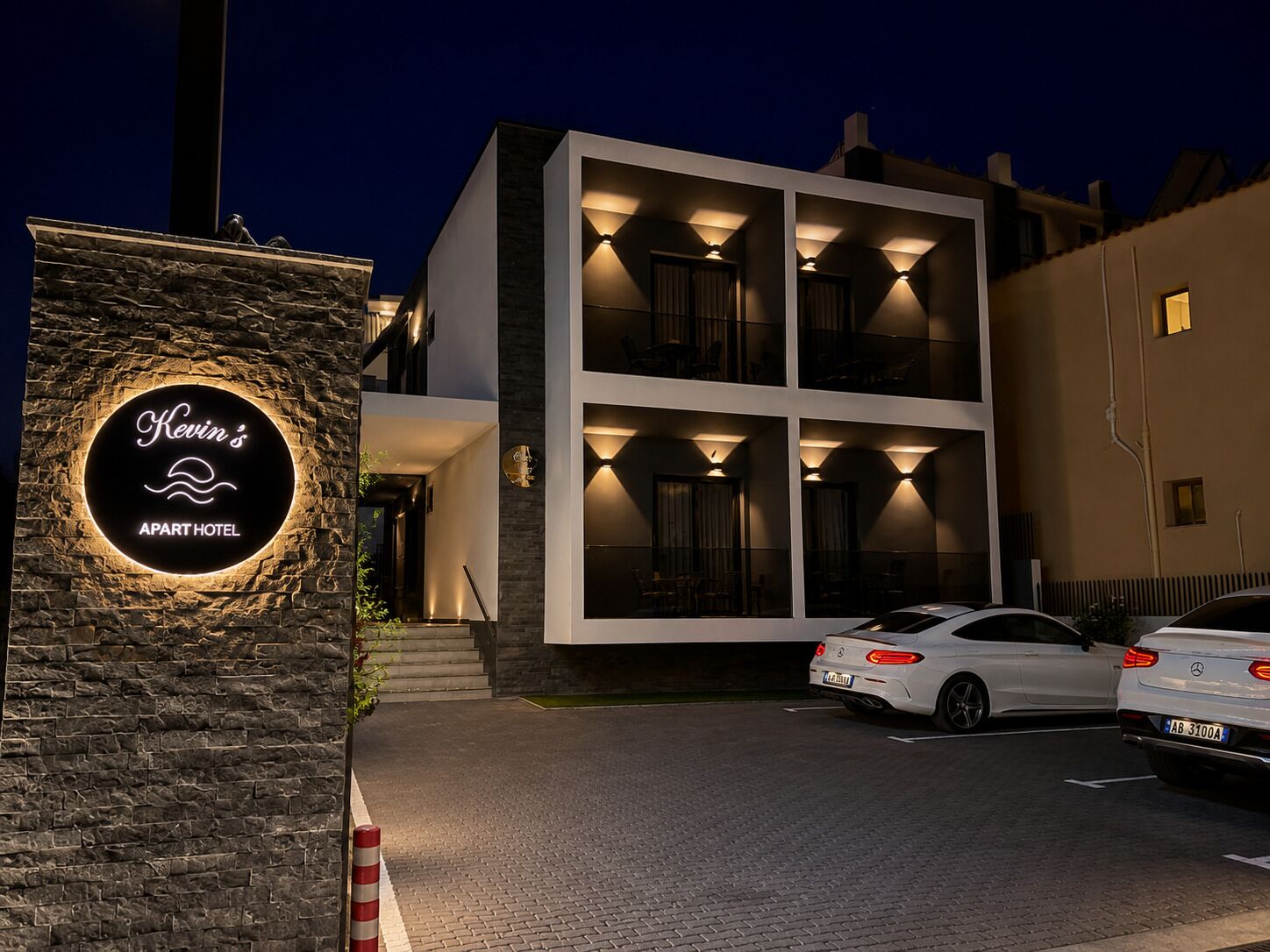Un edificio moderno con un letrero que dice 'Kevin's Apart Hotel' y dos coches estacionados.