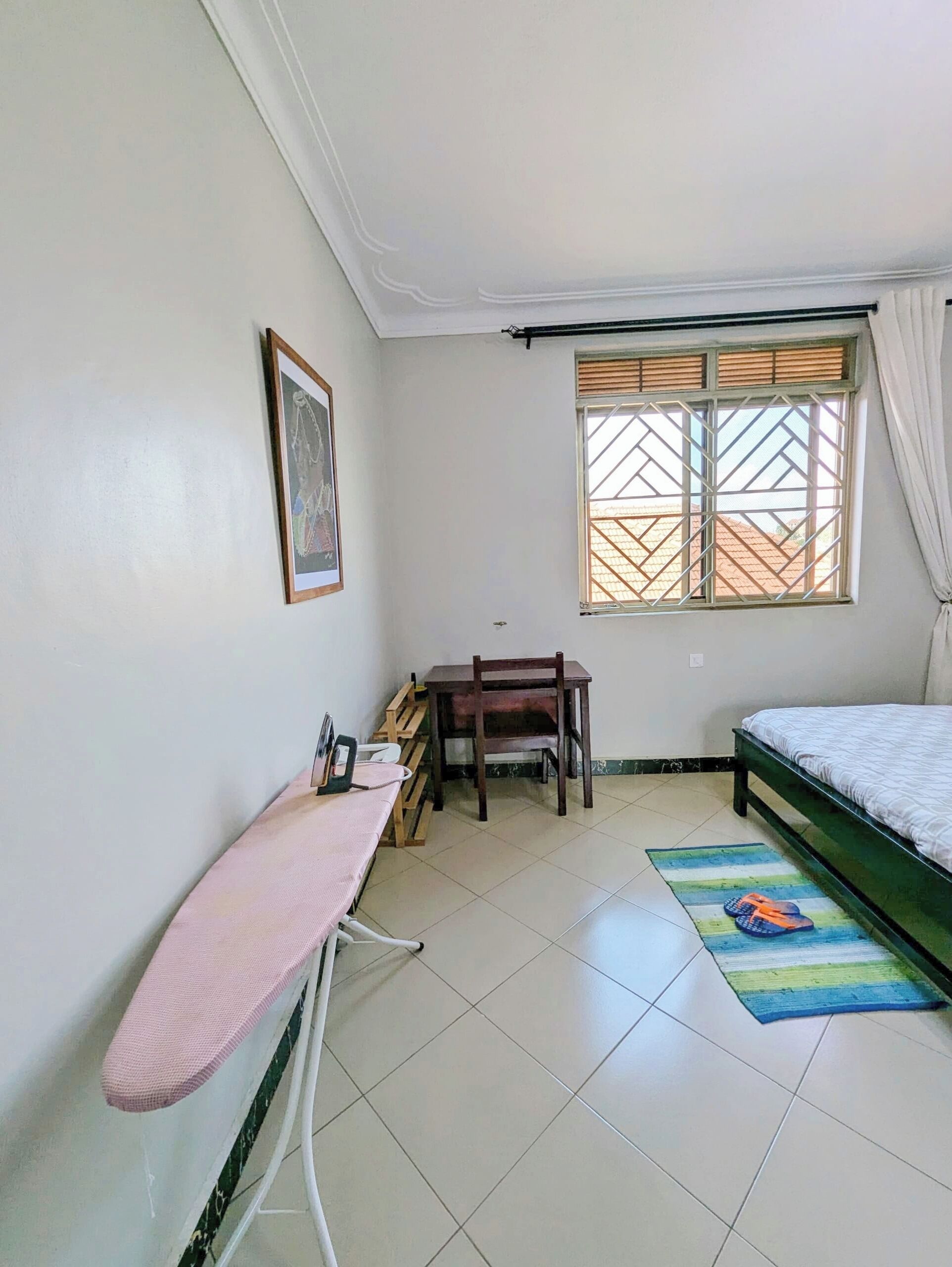 Une chambre à coucher comprenant un lit, un bureau avec des chaises, une fenêtre avec des stores et une planche à repasser fixée au mur.