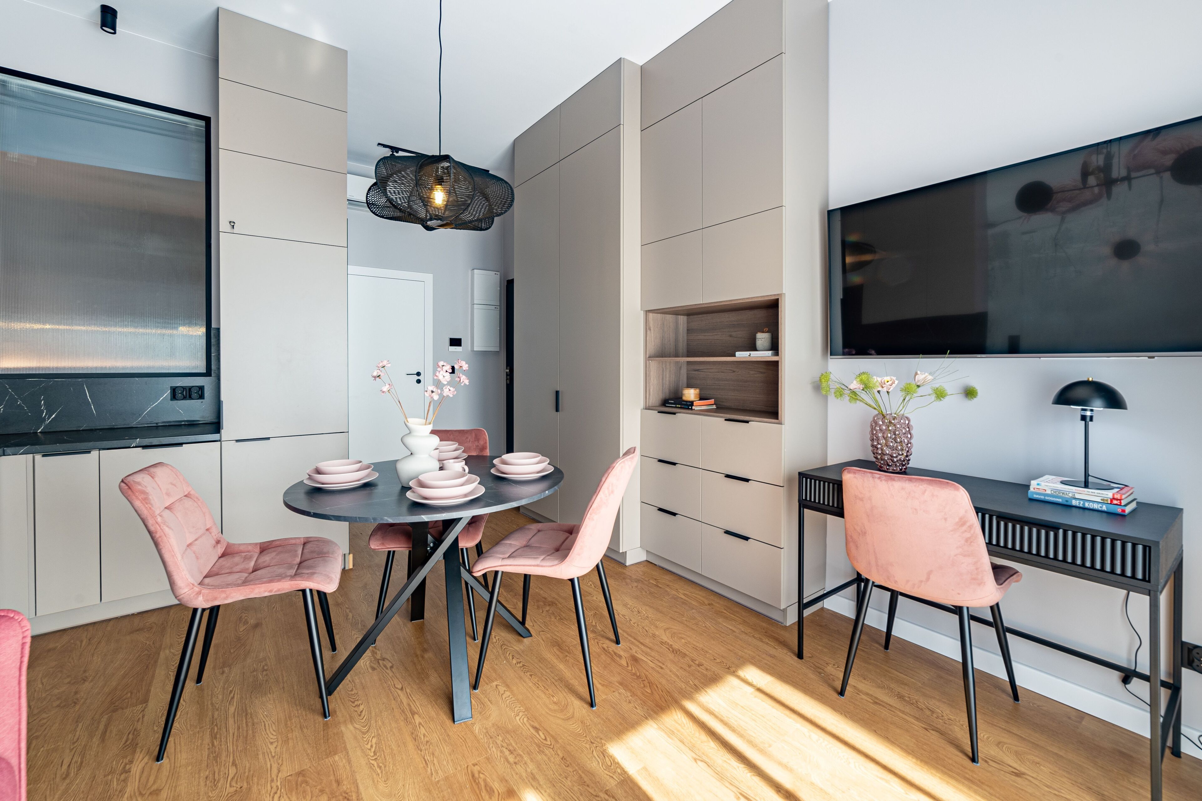 Een moderne keuken met een eethoek, met een ronde tafel, roze stoelen, een zwart bureau en een grote televisie aan de muur.