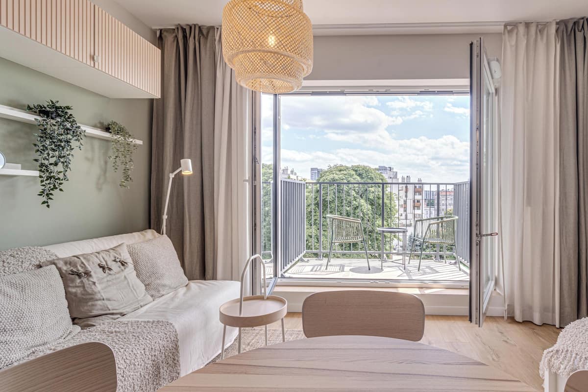 A modern living room with a sofa, a small table, and a chair. There is a balcony with a view of the city and greenery.