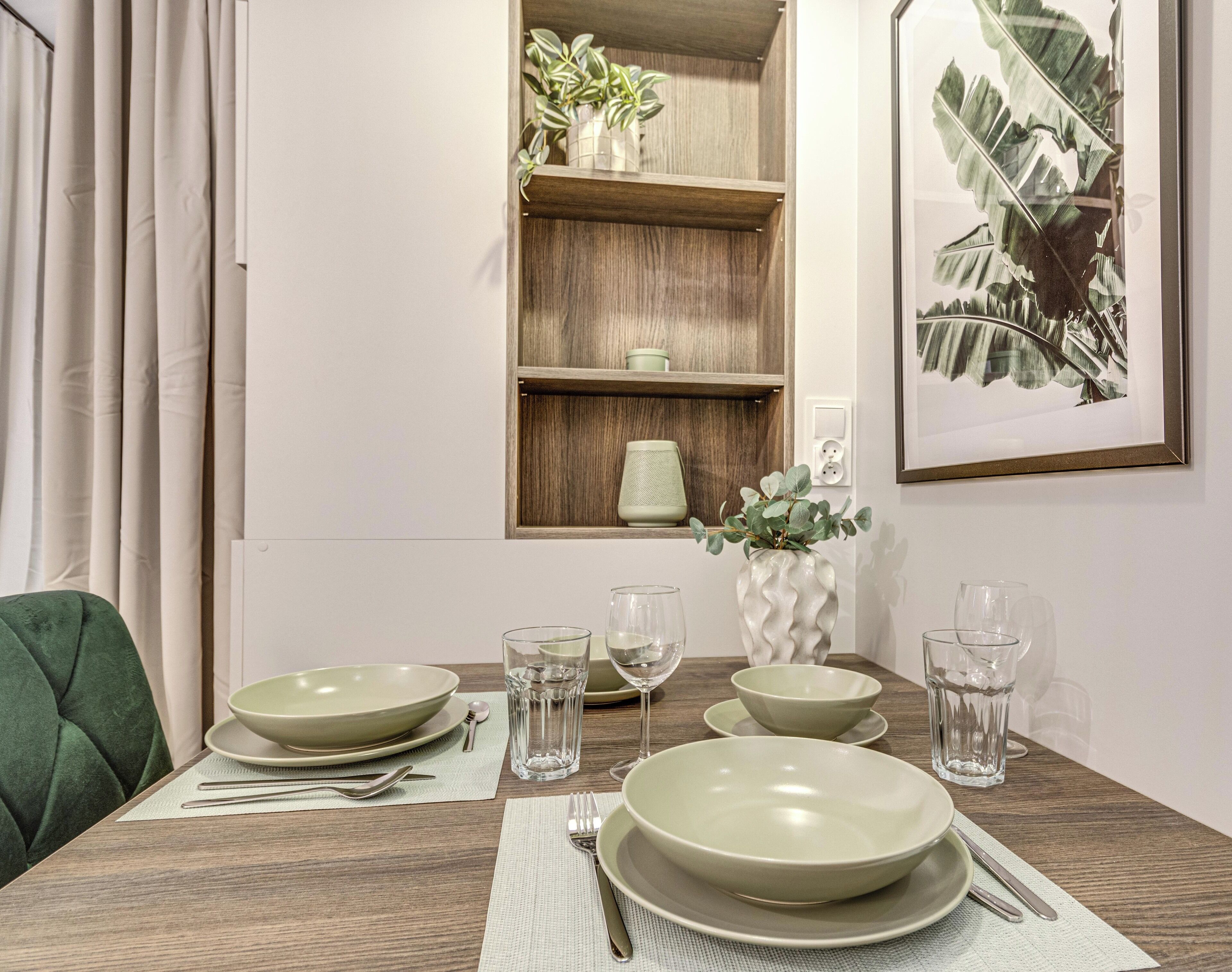 A dining table set with plates, bowls, cutlery, and glasses, with a green chair and a plant on a shelf in the background.