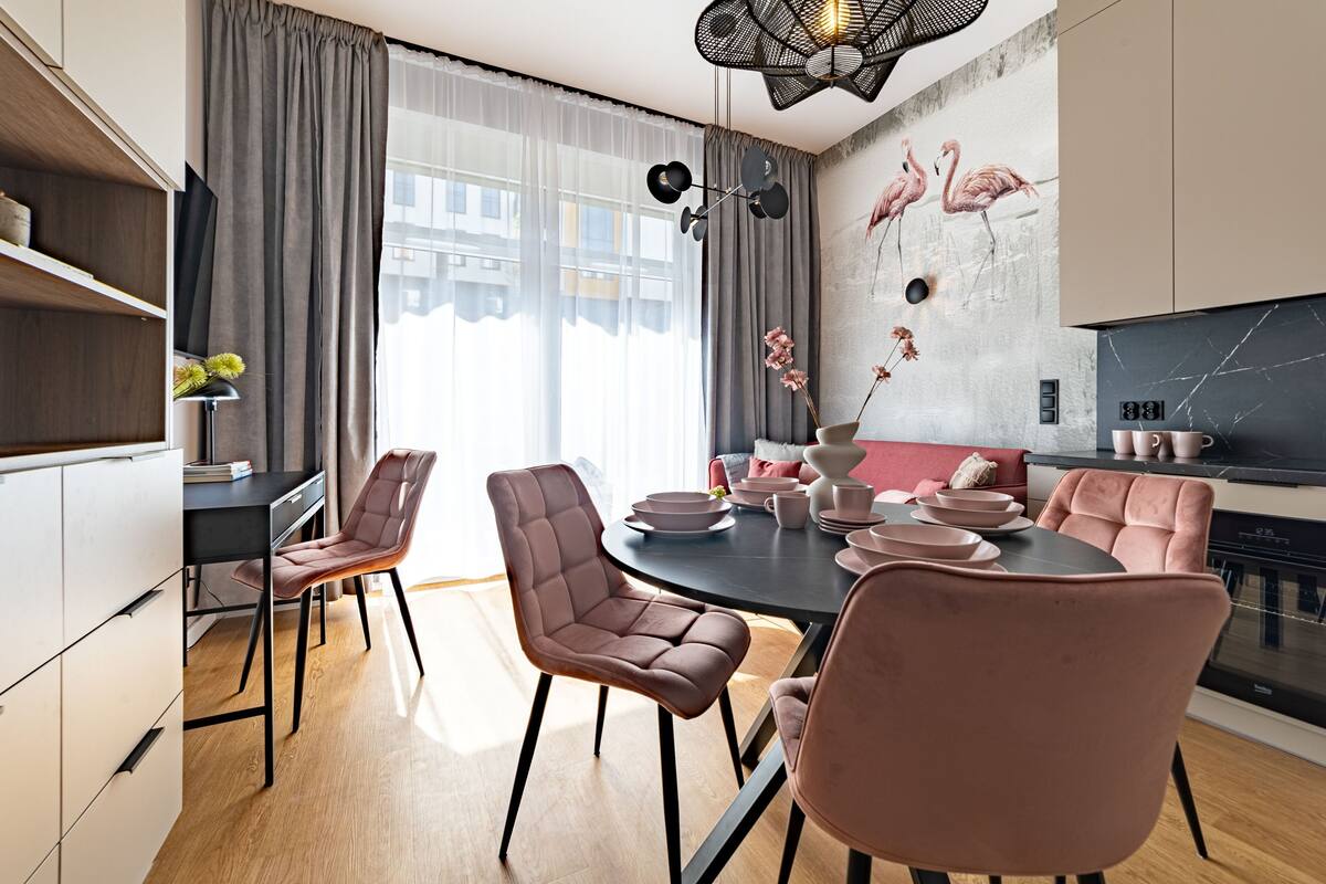 A modern kitchen with a dining area, featuring a black dining table, pink chairs, and a wall with flamingo artwork.