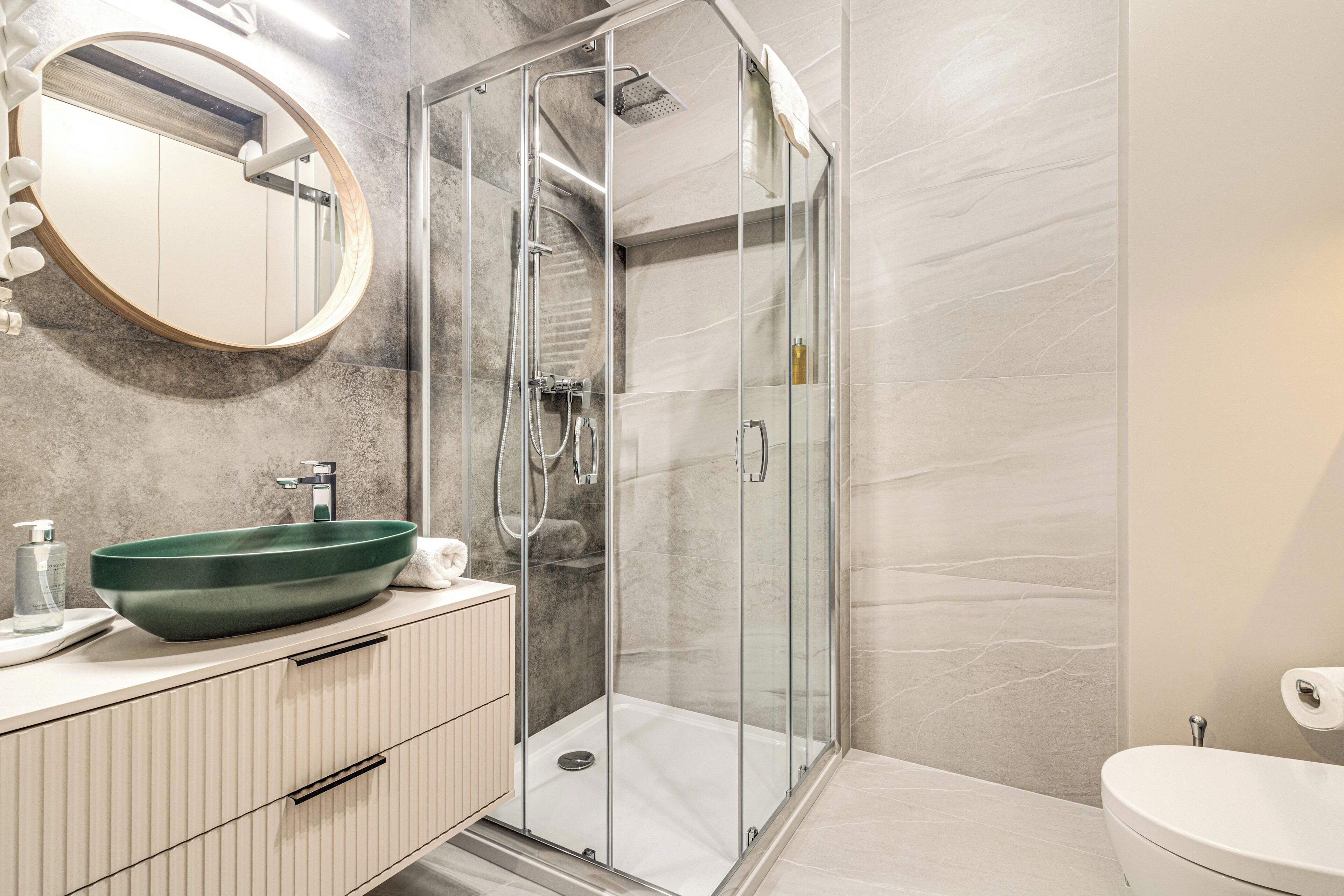 A modern bathroom with a glass-enclosed shower, a round mirror, and a green vessel sink.
