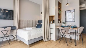 Un dormitorio moderno con cama, escritorio y silla. Hay un televisor en la pared y una ventana con cortinas.