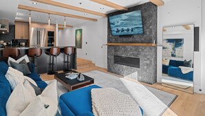 Ein modernes Wohnzimmer mit einem blauen Sofa, einem Couchtisch und einem Kamin. Im Hintergrund ist eine Küche mit Holzschränken und einem Edelstahlkühlschrank zu sehen.