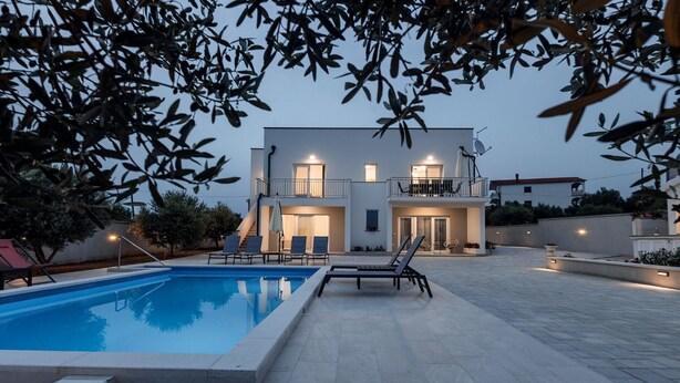 Ein modernes, zweistöckiges Haus mit Swimmingpool, Sitzgelegenheiten im Freien und einem Balkon.