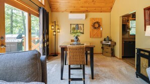 A cozy living room with a dining table, a wicker chair, and a view of the outdoors through large windows.