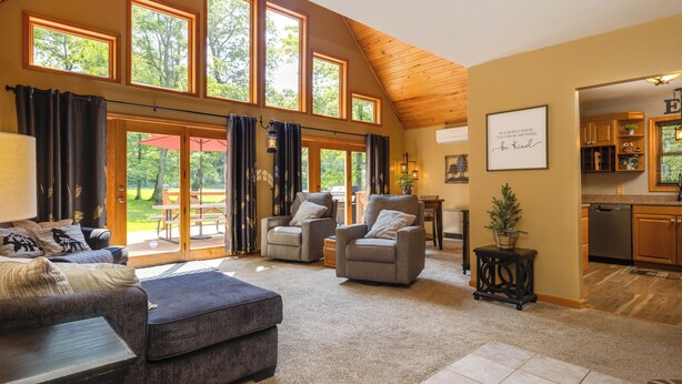 A spacious living room with a high ceiling, large windows, and a view of the outdoors. There are two armchairs, a sofa, and a coffee table. A kitchen is visible in the background.