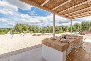 Et udendørs spiseområde med et stentabel og stole, omgivet af en overdækket pergola og kaktusser.