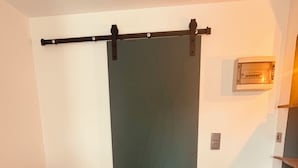 Una habitación con un sofá marrón, un taburete verde pequeño, una puerta corredera y una lámpara fijada a la pared.
