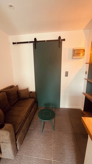 A room with a brown sofa, a small green stool, a sliding door, and a wall-mounted light fixture.