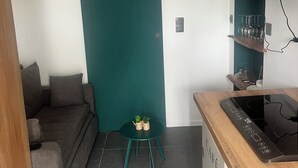 Un espacio habitable compacto con un sofá, una pequeña mesa redonda con dos plantas en macetas, una zona de cocina con estufa y un televisor montado en la pared.