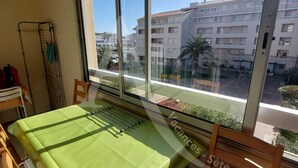 Quarto com uma mesa de jantar coberta por um jogo de mesa verde, uma cadeira e janela com vista para edifícios e uma palmeira.