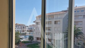 Vista da janela de um prédio residencial com várias varandas, céu claro e rua com carros estacionados e vegetação.