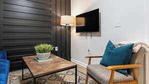 En moderne stue med væg i mørkt træpanel, et fladskærms-tv monteret på væggen, et træsofabord med en plante, en blå sofa og en trælænestol med en turkis pude.