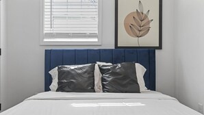 Un dormitorio con cama blanca, cabecera azul y una obra de arte con hojas en la pared.