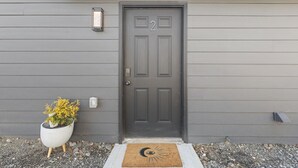 Una puerta gris con el número 2, una planta en maceta y un felpudo con un diseño de luna creciente y un ojo.