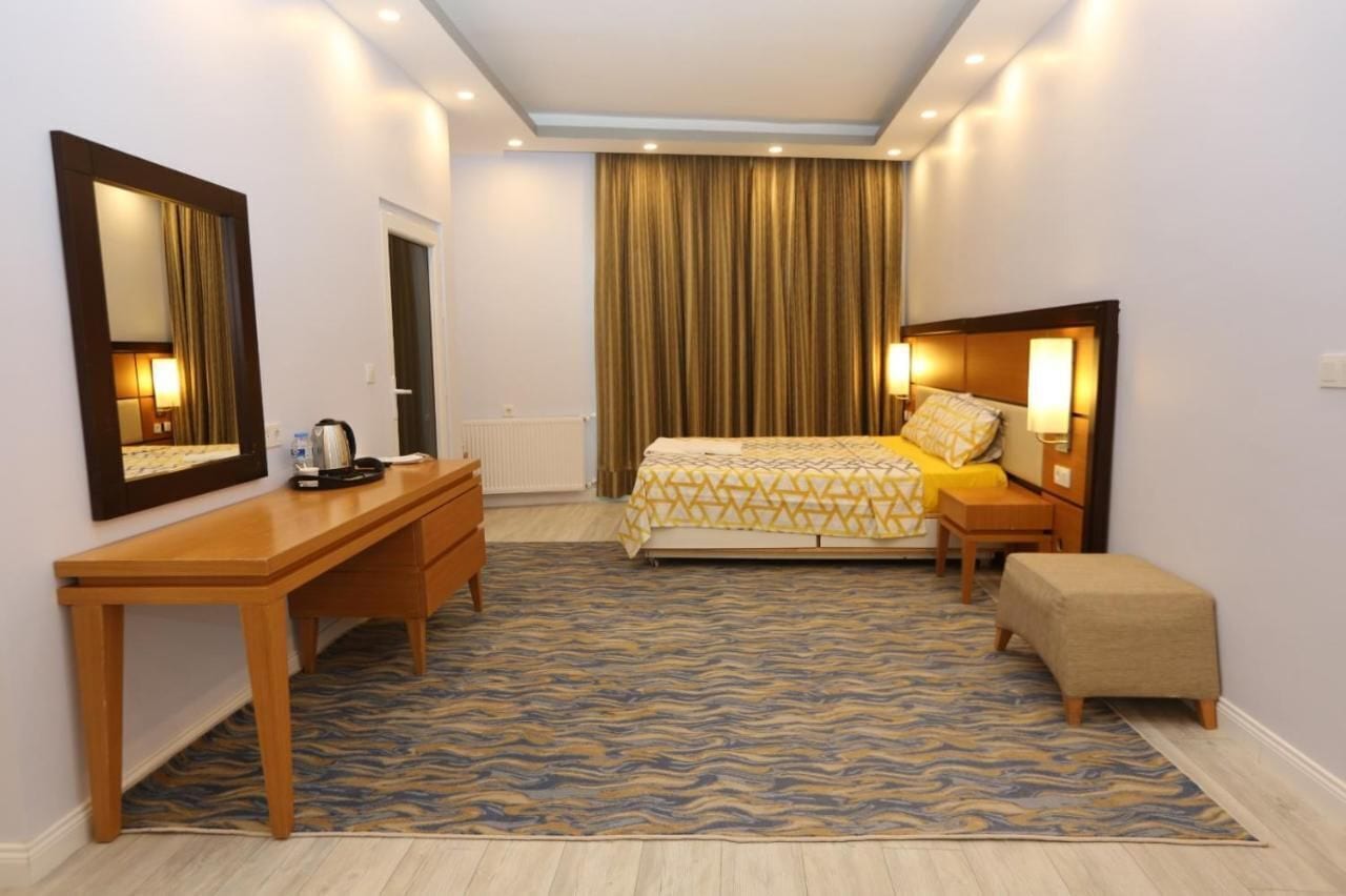 A hotel room with a bed, a desk, a chair, a bench, a mirror, and a curtain.