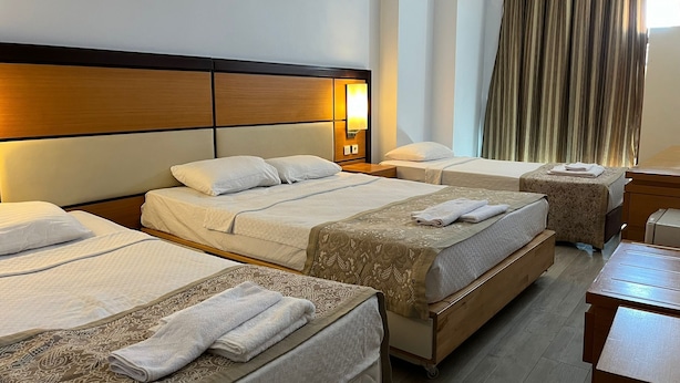 A hotel room with three beds, a wooden headboard, a nightstand, a desk, and a large window with curtains. - Kommageneiz Hotel (Kahta)