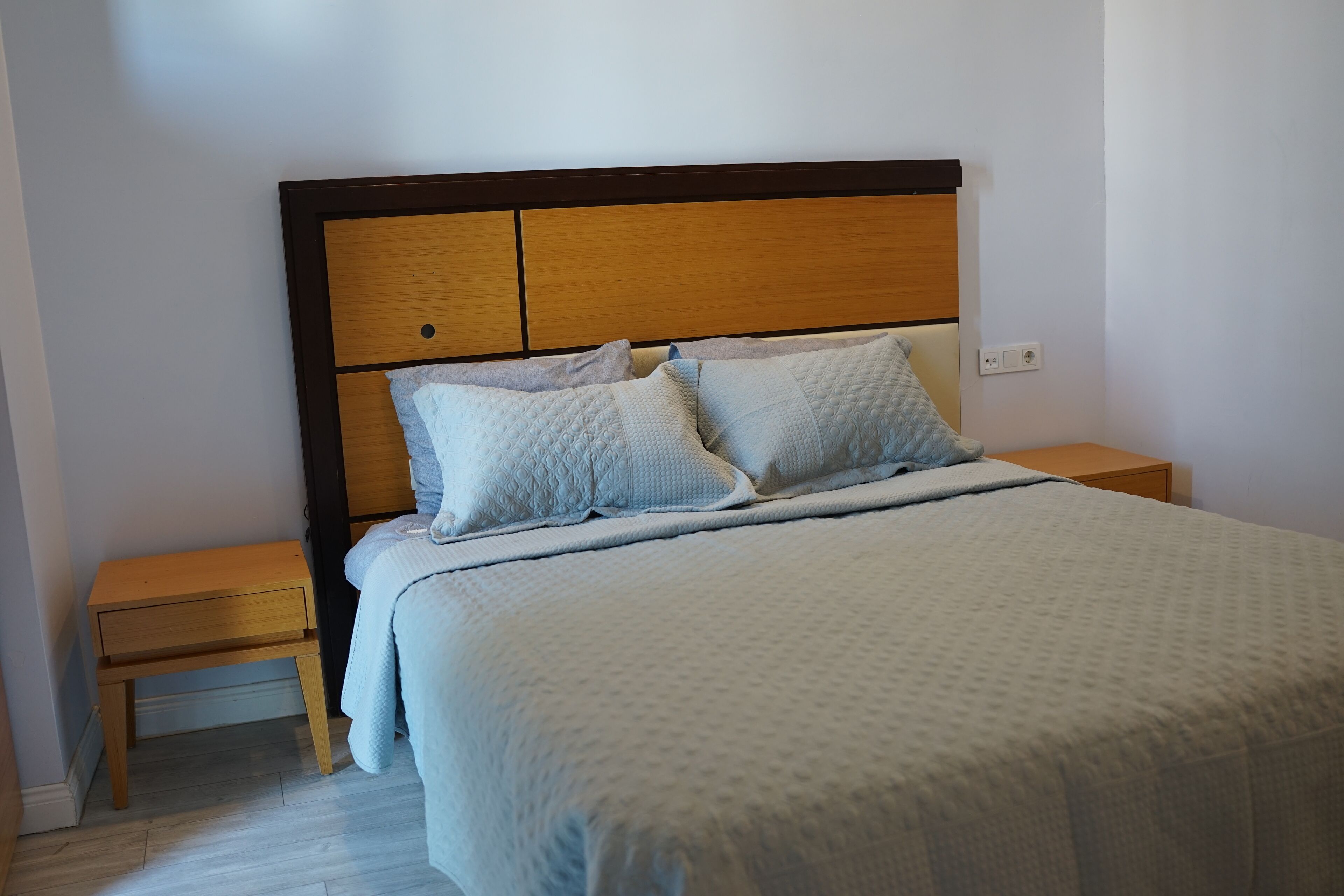 Una cama bien tendida con cabecera de madera, una mesita de noche de madera a un lado y suelo de color claro.