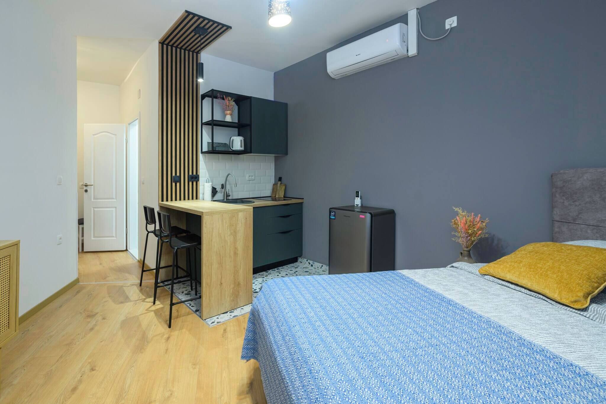 Habitación pequeña con cama, una kitchenette, un escritorio con sillas y una puerta que da a otra habitación.