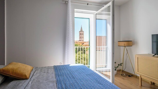 A bedroom with a bed, a window with a view of a building, a floor lamp, and a wooden cabinet. - Vera Studio Deluxe With Oldtown View (Zadar)