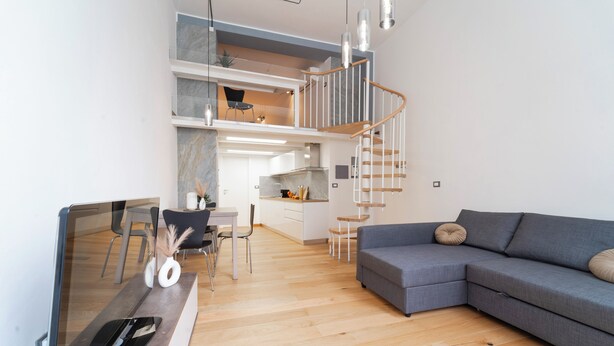 A modern living room with a grey sectional sofa, a dining area with a table and chairs, a kitchen in the background, and a spiral staircase leading to an upper level. - Milan Lambrate M2 Center (Milano)
