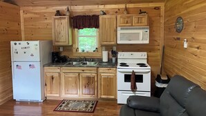 A compact kitchen with wooden cabinets, a refrigerator, a microwave, a stove, and a black leather couch. - Lowry’s Retreat (South Bloomingville)