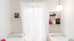 Een minimalistisch ingerichte slaapkamer met twee enkele bedden, een groot raam met dunne gordijnen, een nachtkastje met boeken en een plant, en een hanglamp.