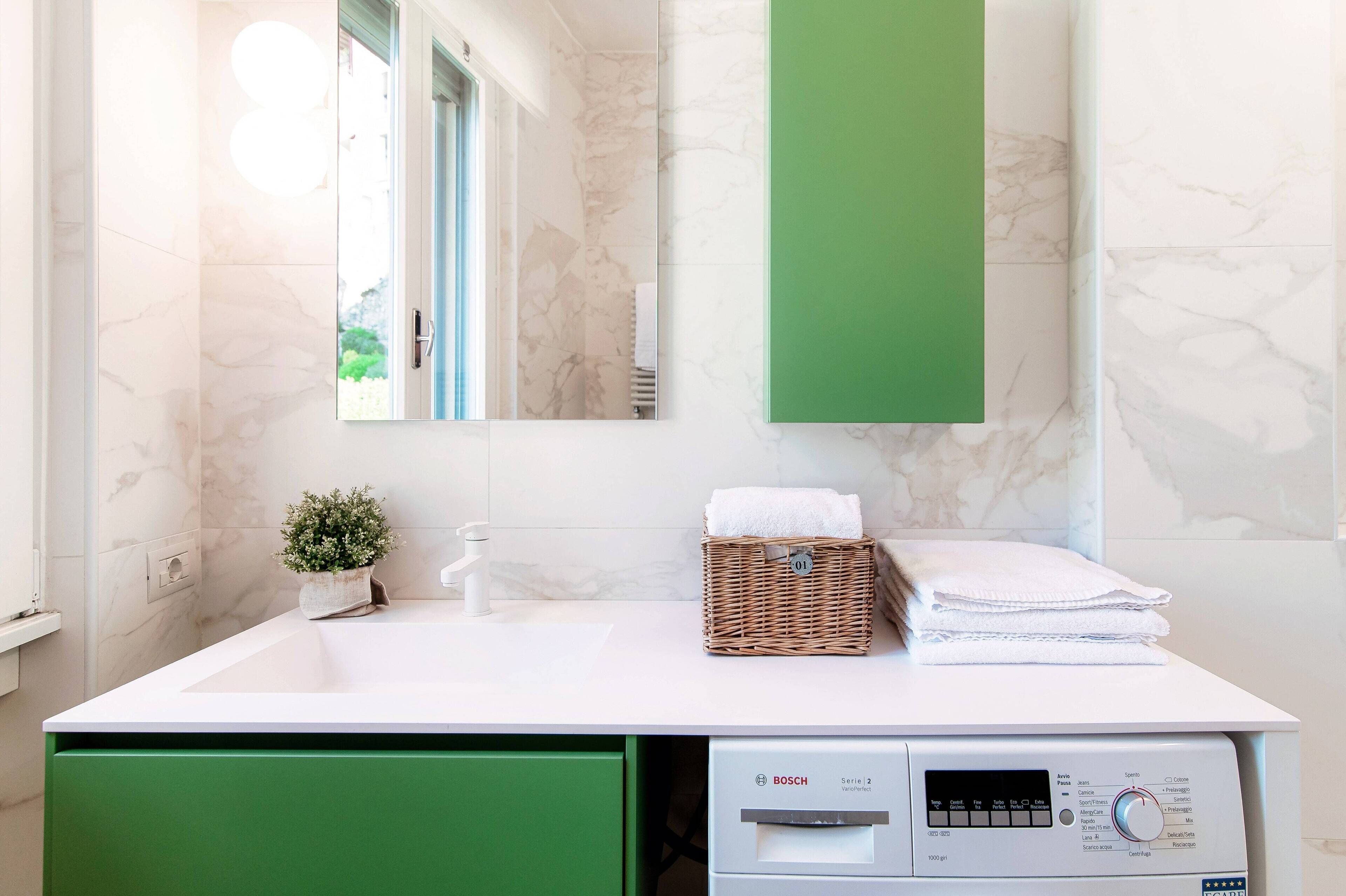 A modern bathroom with a white sink, a green cabinet, a Bosch washing machine, and a mirror with a green accent wall.