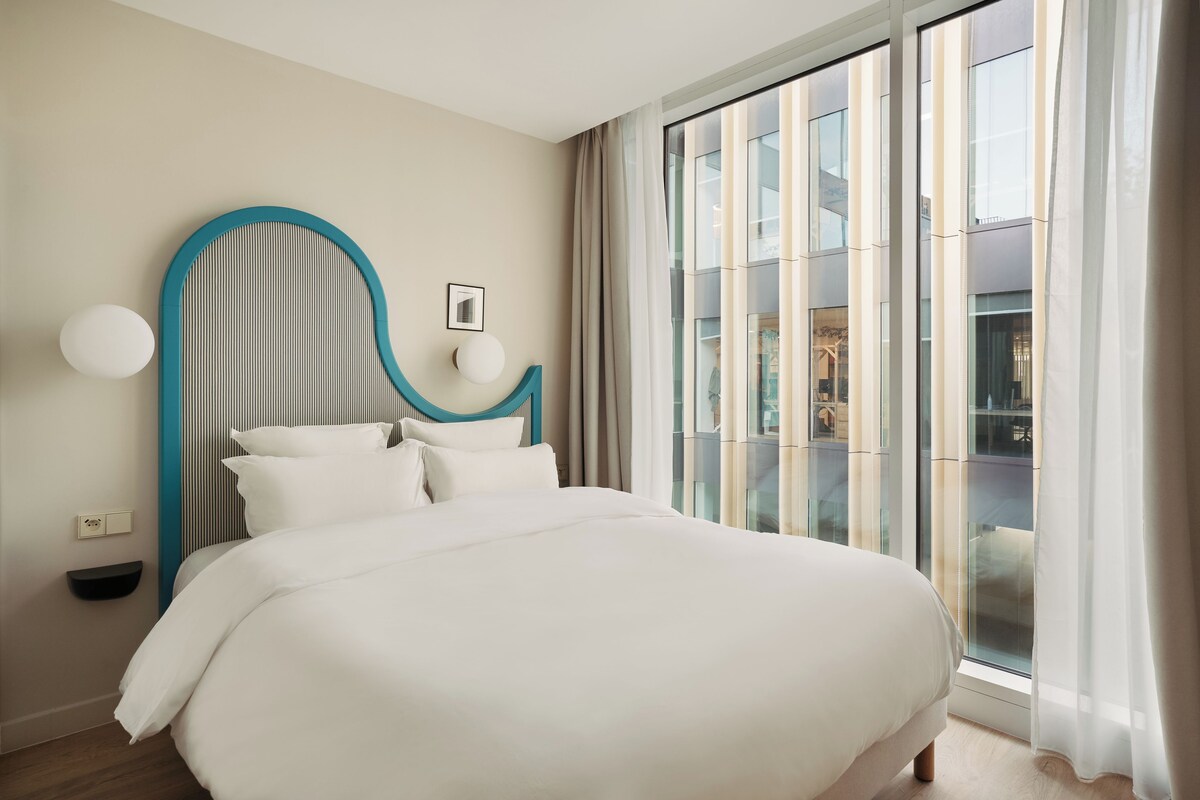 A modern bedroom with a large bed, a headboard with vertical stripes, a bedside table, and a view of a building through large windows.