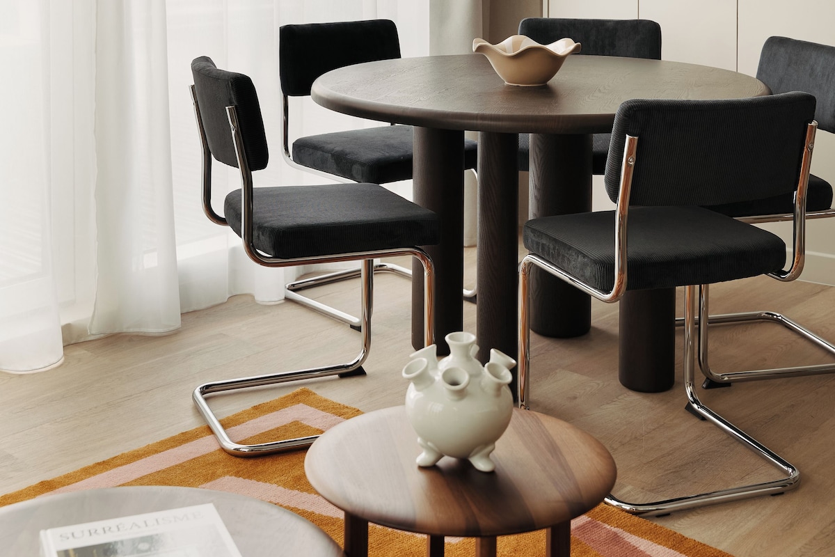 A modern dining area with a round table, black upholstered chairs, a pendant light, and a white vase on the floor.