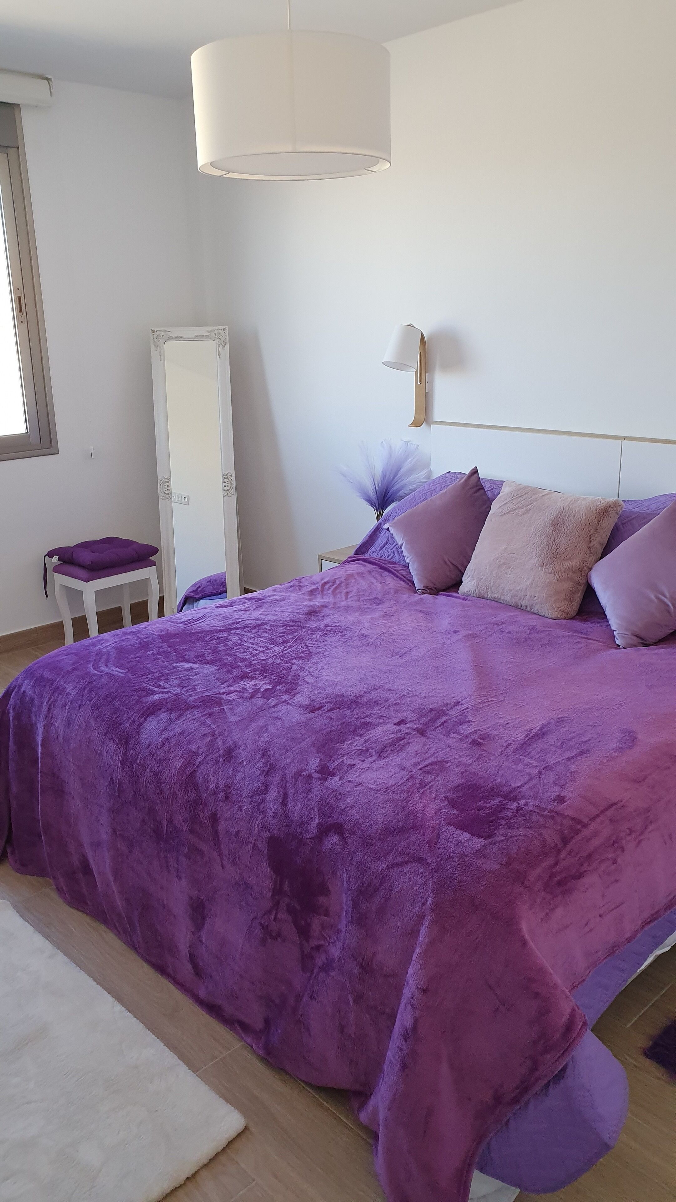 A bedroom with a large bed, a bedside table, a chair, a mirror, and a hanging light fixture.