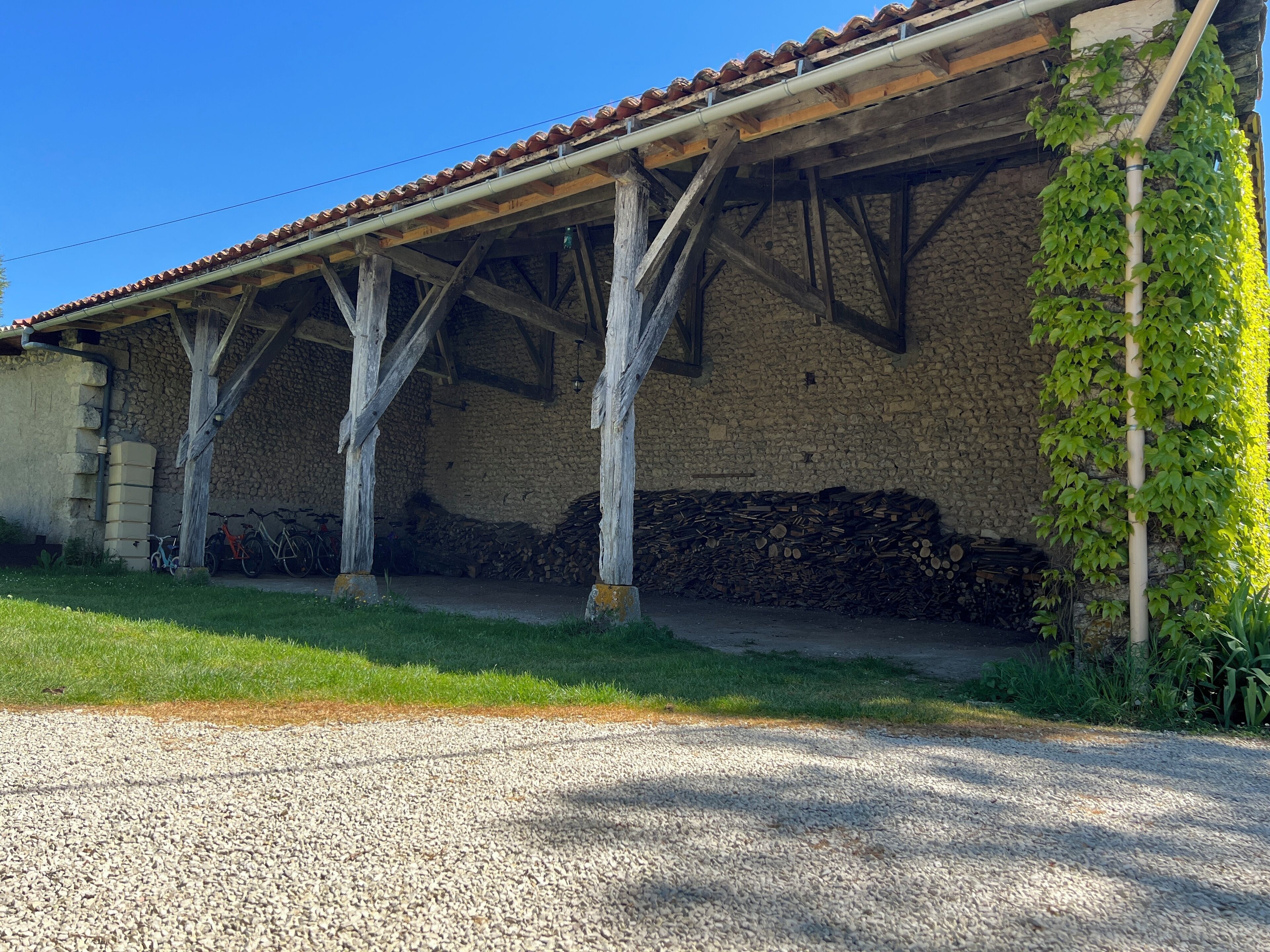 Ein Gebäude mit einer Steinwand und einem Ziegeldach. Holzträger stützen das Dach, und ein Holzstapel lehnt an der Wand.