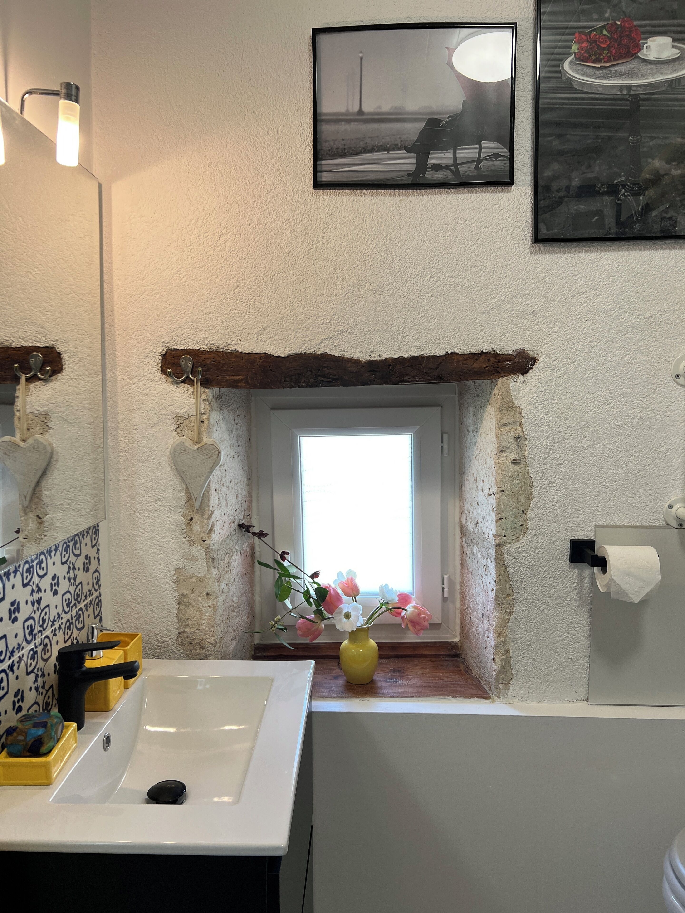 A bathroom with a white sink, a yellow vase with flowers, a framed picture above the mirror, and a window with a view of a lamp post.