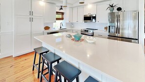A modern kitchen with white cabinetry, stainless steel appliances, and a large island with bar stools. - Oceanside Unit A | Luxe Beachfront! (Tybee Island)