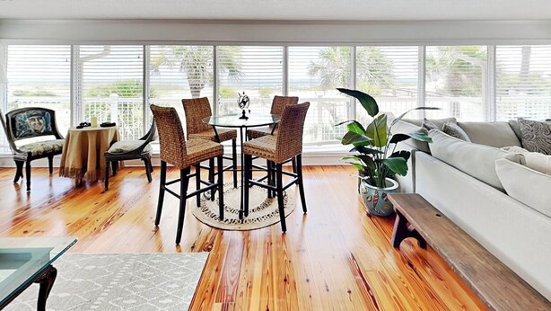 A spacious living room with a wooden floor, a round table with chairs, a sofa, and large windows with blinds. - Oceanside Unit A | Luxe Beachfront! (Tybee Island)