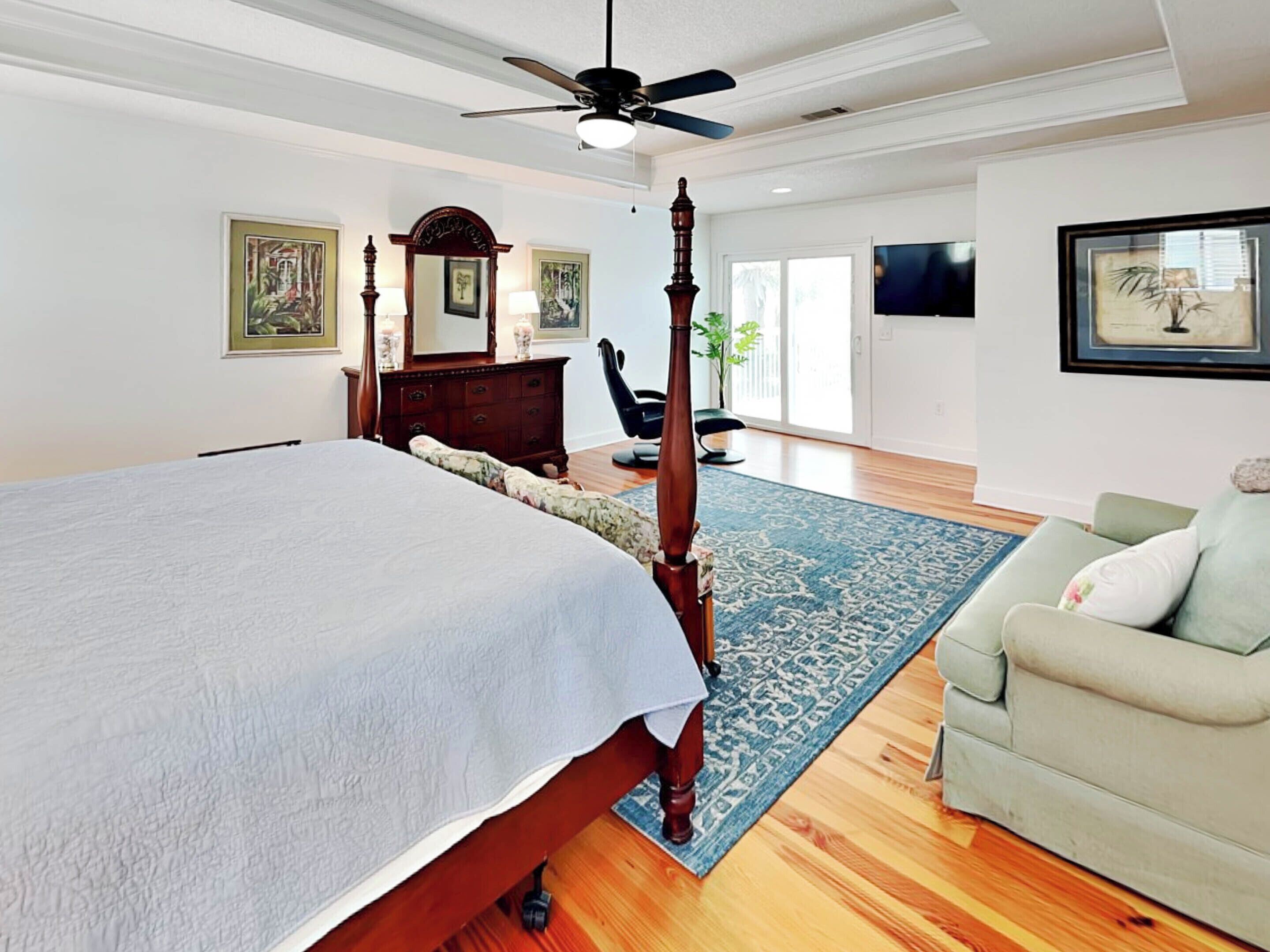 A bedroom with a large bed, a ceiling fan, a wooden dresser, a chair, and a television.