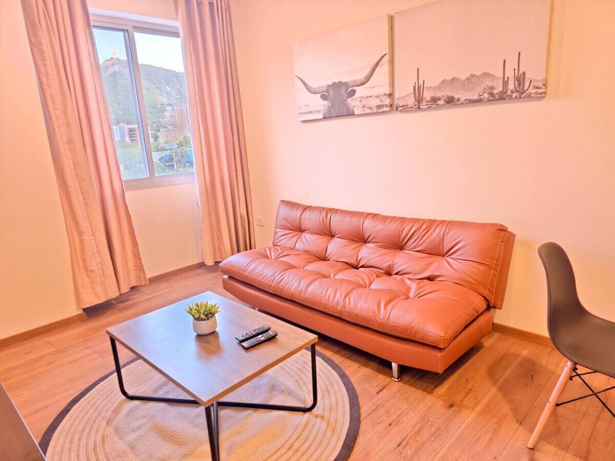 Una sala de estar con un sofá de cuero, una mesa de centro con una planta, un control remoto y un cuadro con un toro y cactus en la pared.
