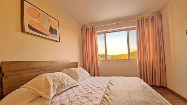 A bedroom with a large bed, a wooden headboard, a window with curtains, and a painting on the wall. - Modern apartment in Recoleta (Cochabamba)