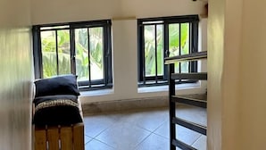 A room with a high ceiling, three framed black-and-white photographs on the wall, a wooden bench with a black cushion, and two windows with a view of greenery. - Sawe Homes 4&5 (Kampala)