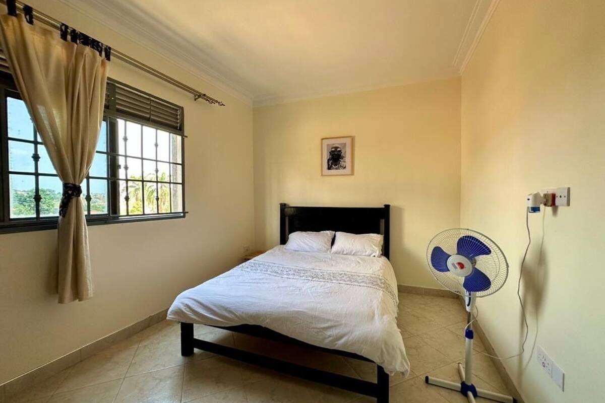 Une chambre à coucher avec un lit, une fenêtre avec des rideaux, un ventilateur fixé au mur et un tableau encadré.