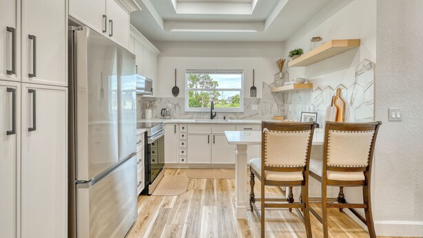 A modern kitchen with white cabinets, a stainless steel refrigerator, a window, and a dining area with two wooden chairs. - Charming 3-bedroom Relaxing Family Vacation Retreat On The Water,  Fishing (Rotonda West)