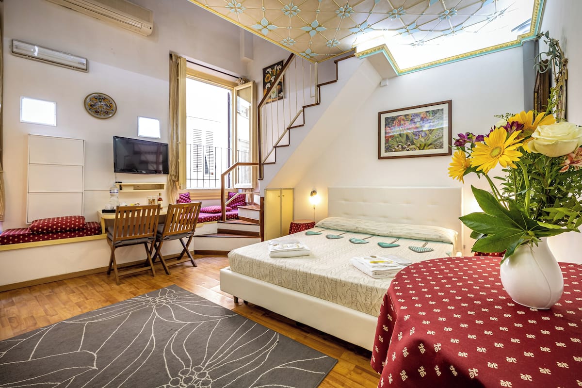 A bedroom with a bed, a desk, a chair, a TV, a window, and a staircase.