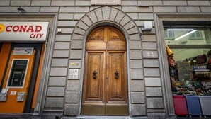 En byggnadsfasad med en stor, dubbel träport, en skylt med texten ”CONAD CITY” och ett fönster där resväskor visas.