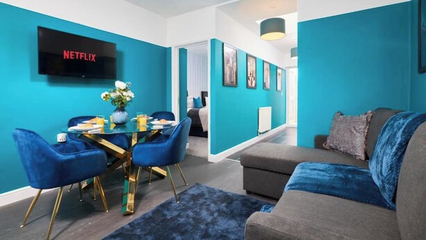 Una sala de estar moderna con una pared azul, un televisor de pantalla plana, una mesa de comedor, sillas azules y un sofá gris.