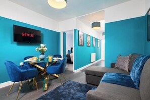 Una sala de estar moderna con una pared azul, un televisor de pantalla plana, una mesa de comedor, sillas azules y un sofá gris.