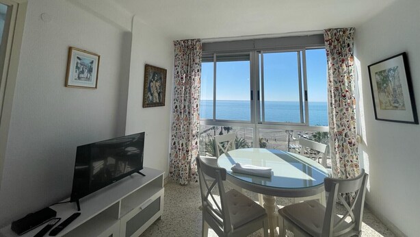 A room with a TV on a stand, a glass table with chairs, and a view of the sea through large windows. - TERRASOL FRONTAL AL MAR ANTILLAS 8 (Torre del Mar)